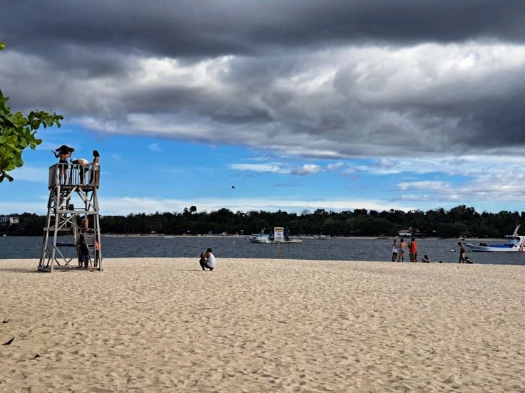 Sandy beach with a lifeguard tower, scattered visitors, and boats under a cloudy sky.