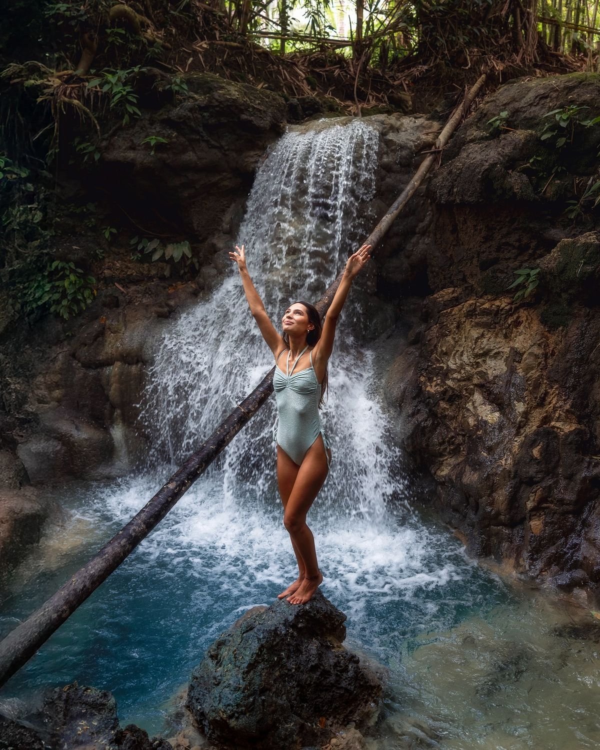 Woman standing on a rock beneath a jungle waterfall with arms raised.