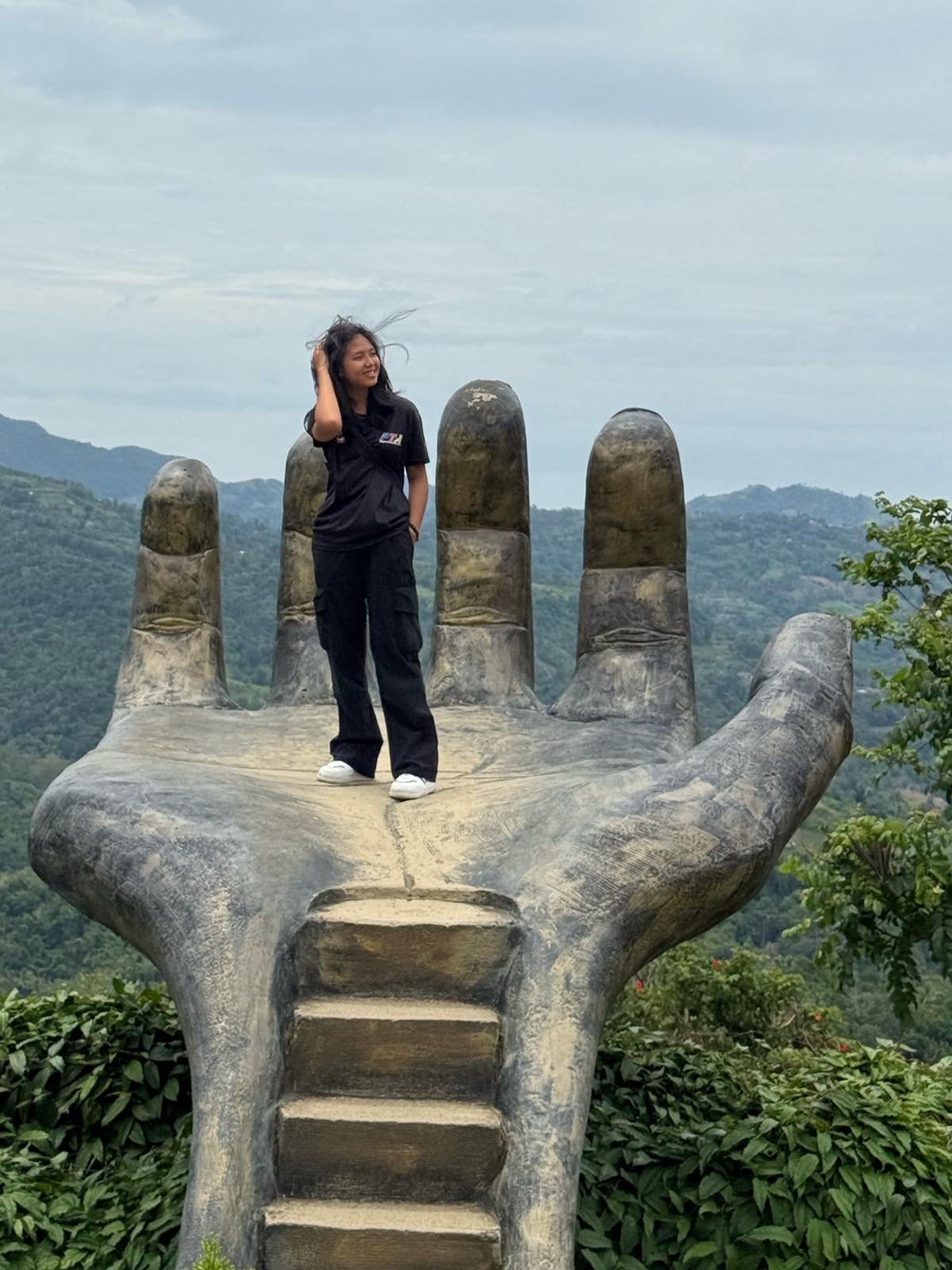 Home A traveler posing on the iconic giant hand sculpture at a Cebu highland attraction, a highlight of the customized Cebu City mountain tours curated by Cebu Travels & Oslob Tours.