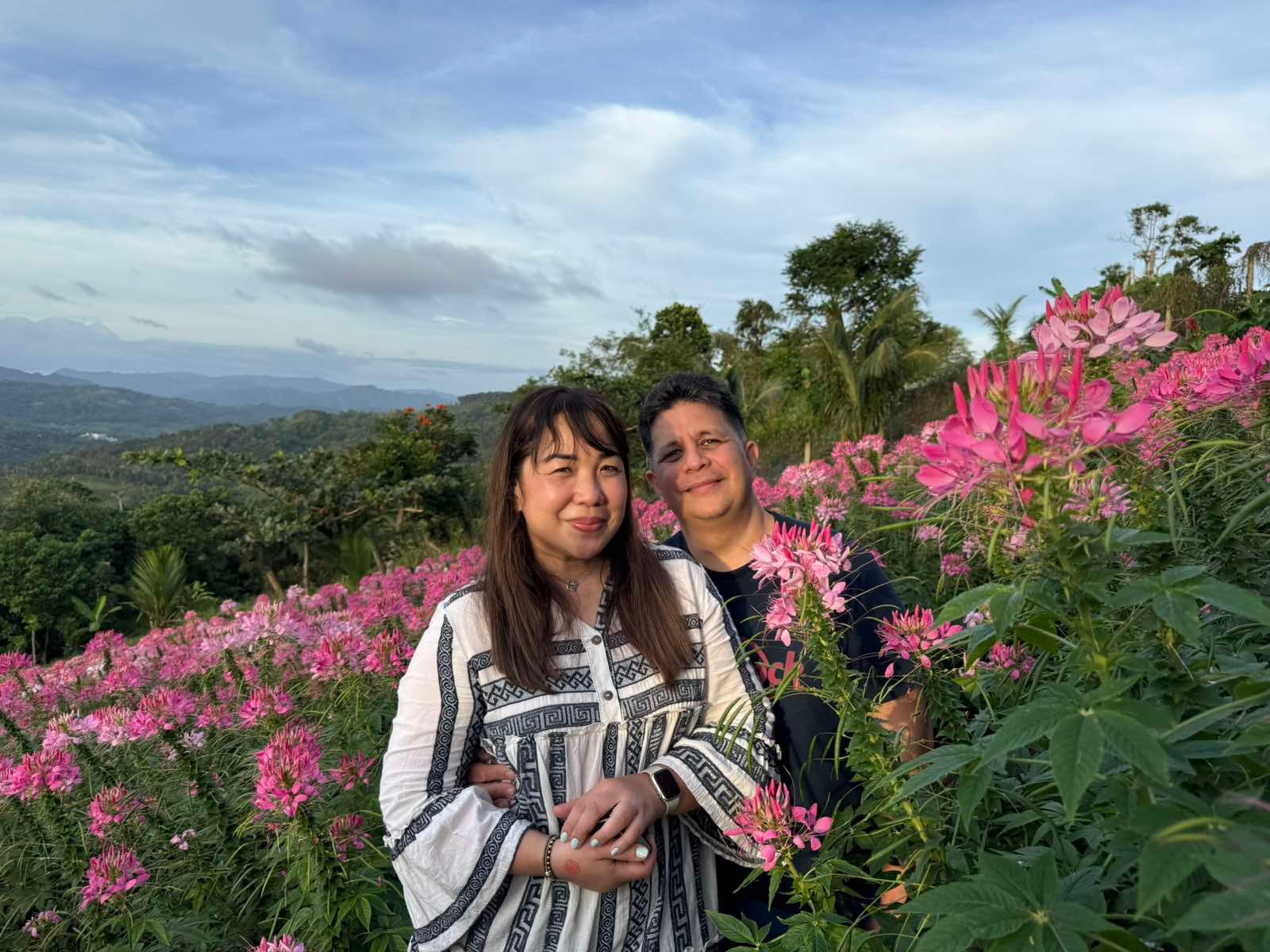 Home A couple poses together in a vibrant field of pink flowers on a hillside during a scenic mountain tour with Cebu Travel & Oslob Tours.