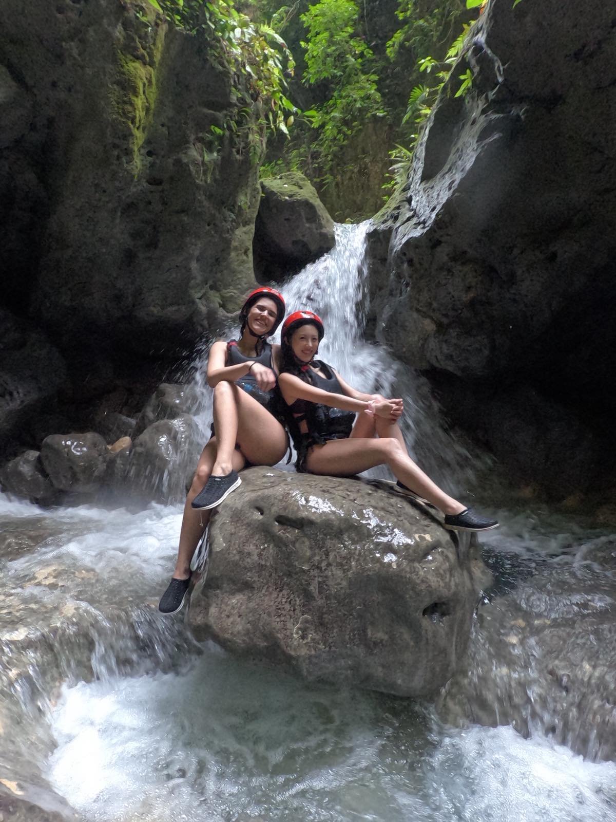 Home Two travelers wearing red safety helmets and life vests sit on a large rock in the middle of a flowing river with a small waterfall in the background during a canyoneering adventure in Badian, Cebu.